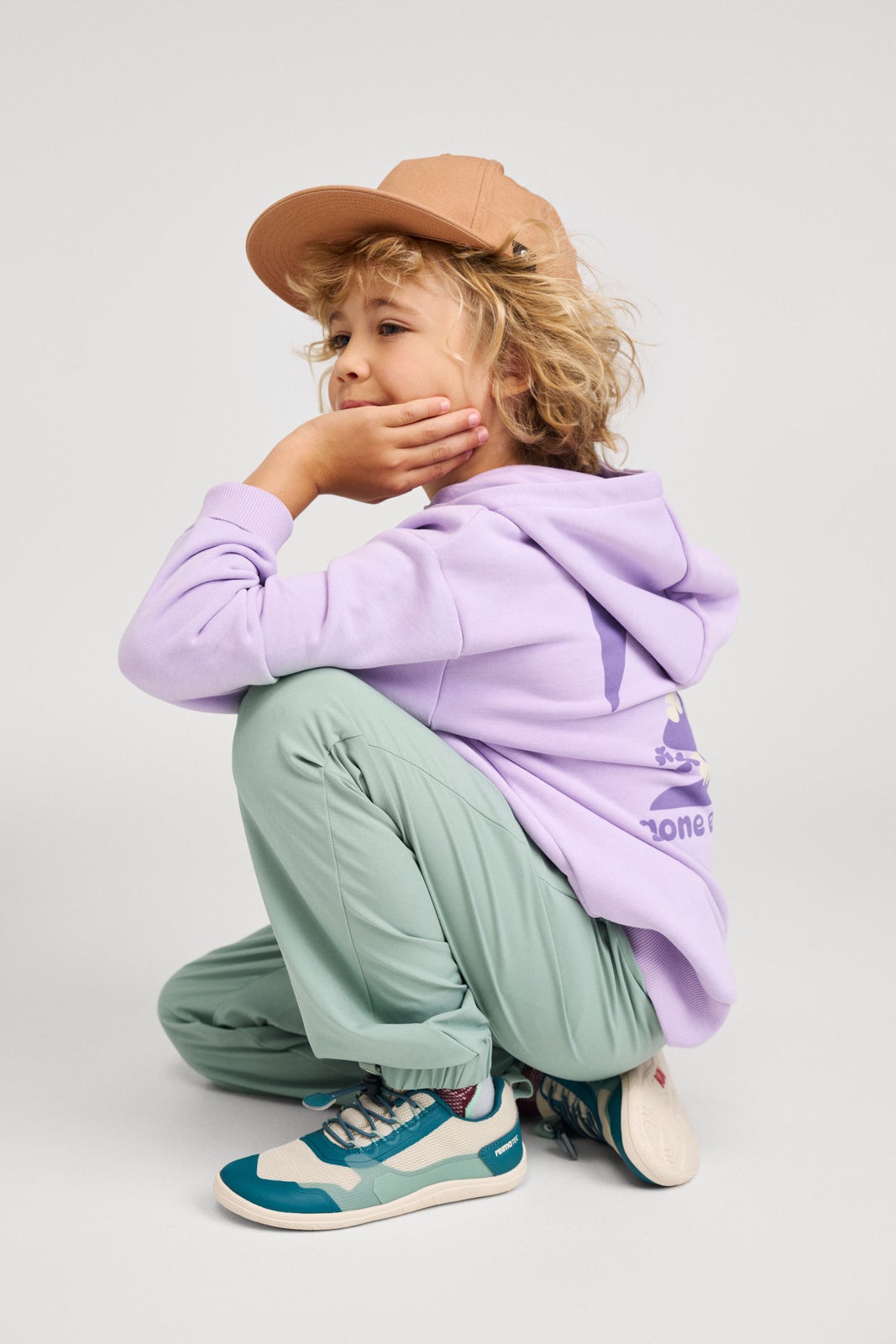 Lifestyle shot of a child wearing the blooming lilac Reima Huppis organic cotton hoodie in a crouching pose, paired with green outdoor pants, a tan cap, and sneakers, demonstrating its flexible, active fit for everyday play.