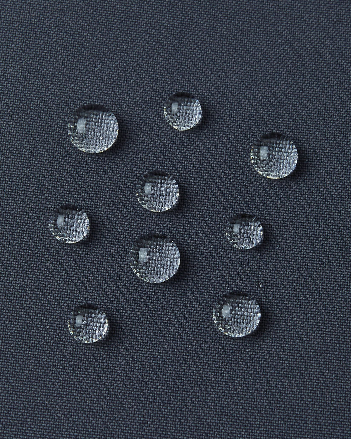 A detailed close-up texture shot of several perfectly formed water droplets beading up cleanly on the dark surface of the water-repellent fabric of a Reima Kuosku kids ski jacket (Soft black color), demonstrating its durable, fluorocarbon-free performance.
