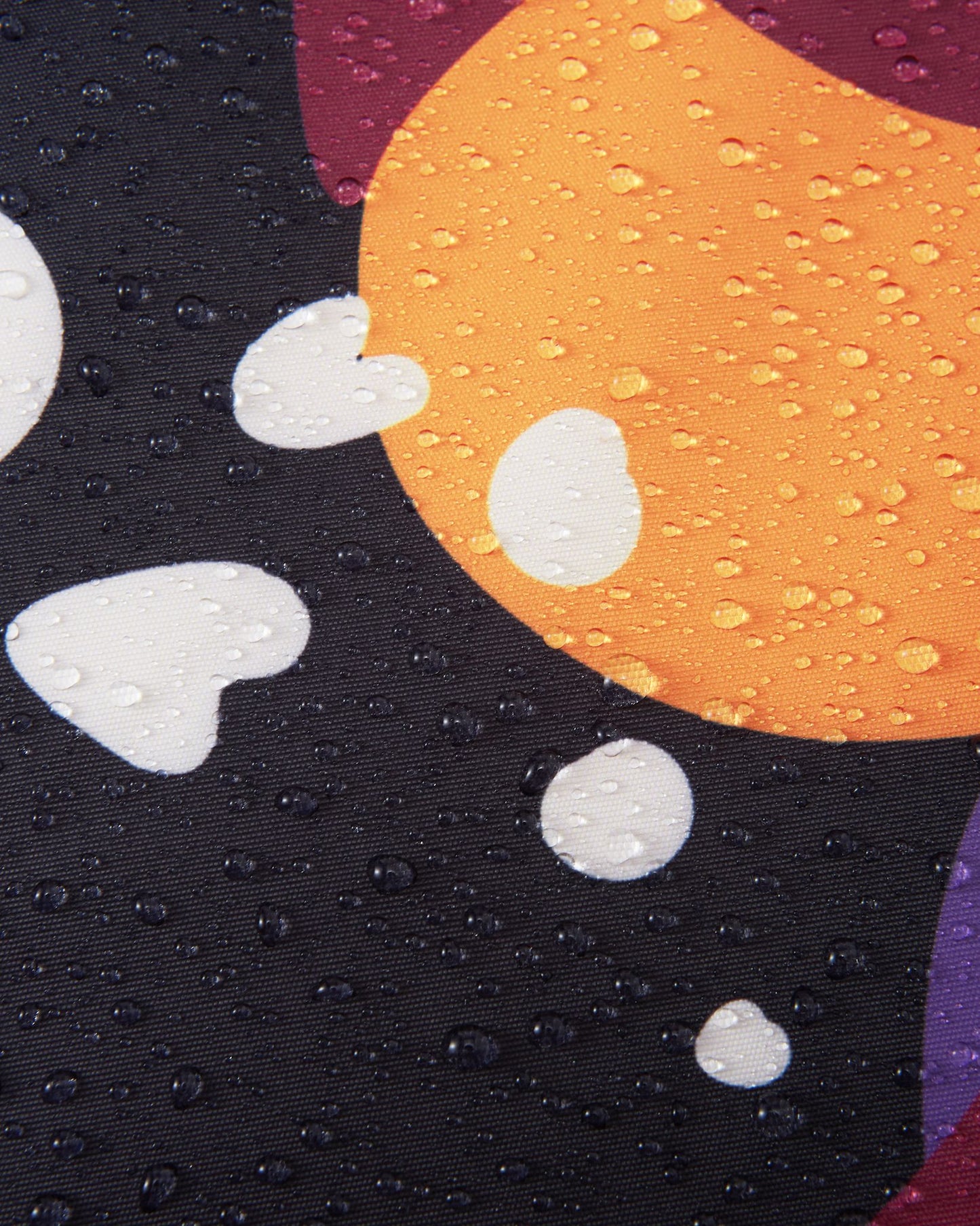 Macro close-up of water droplets tightly beading on the colorful black, orange, and purple Reimatec fabric of the Reach ski suit, highlighting its exceptional waterproof and weather-resistant performance in wet, snowy conditions.