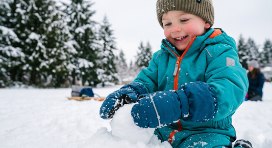 A toddler in a teal snowsuit, a knit beanie, and large navy blue mittens forms a snowball in the snow against a snowy evergreen forest background.
