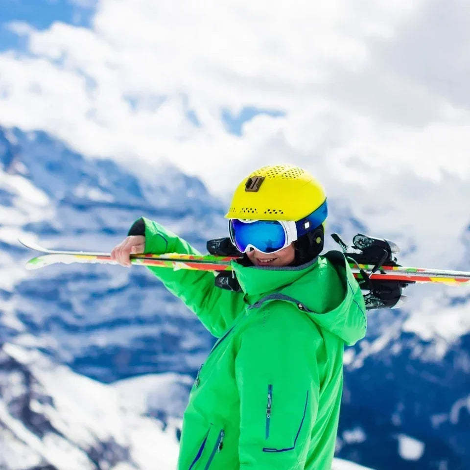 A child wearing a green winter jacket, yellow helmet, and blue reflective kids' ski goggles carrying skis over their shoulder on a sunny, snow-covered mountain, demonstrating the need for UV protection against high-altitude sun glare.