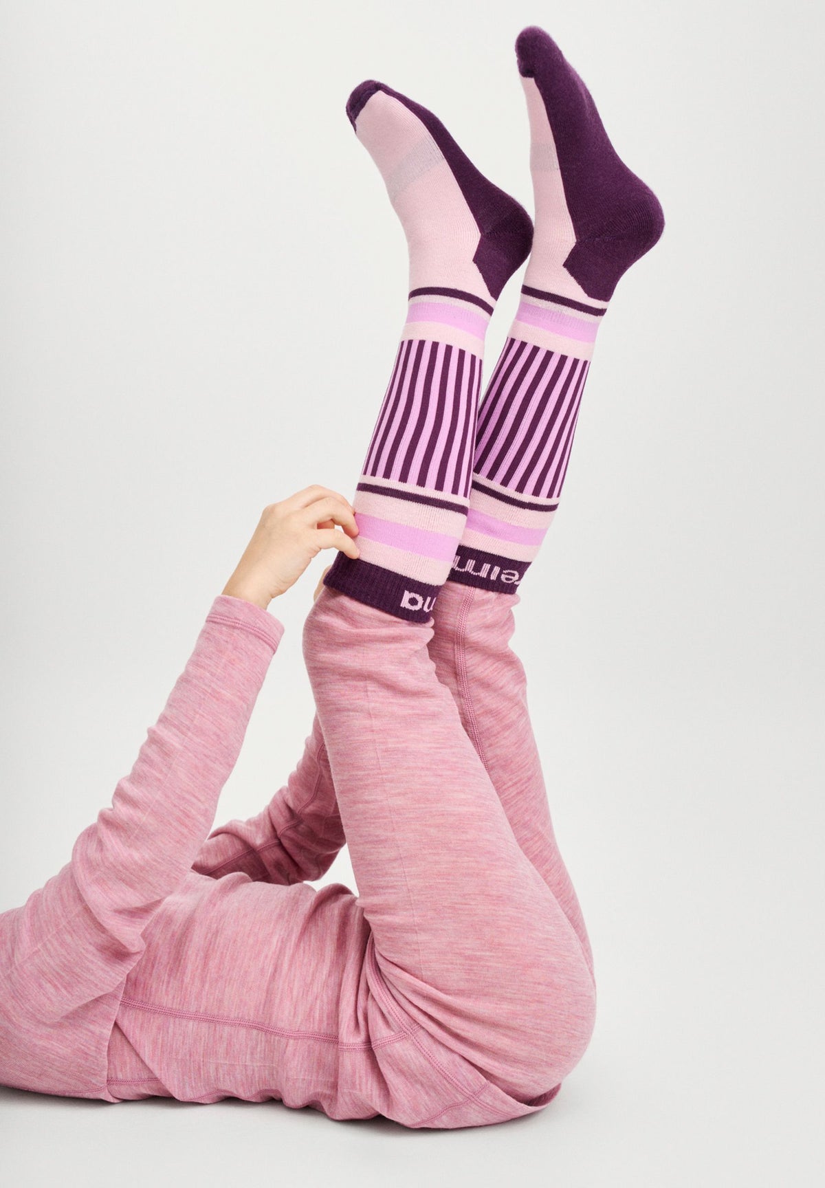 A child lying comfortably on their back with their legs raised in the air, wearing the grey pink Reima Kinsei merino wool base layer set paired with thick purple and pink Reima thermal ski socks.