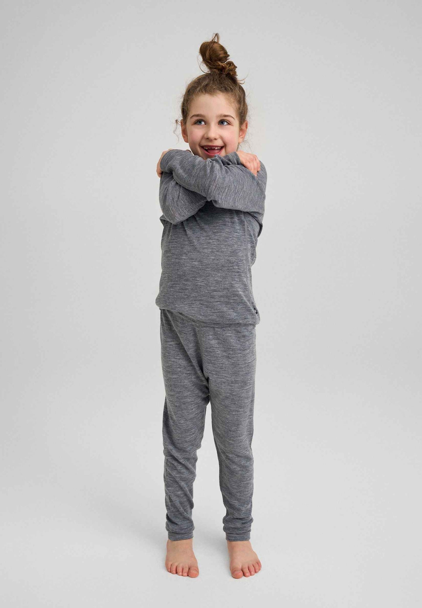 A happy child hugging herself to stay cozy while wearing the full Reima Kinsei kids' merino wool base layer set in melange grey.