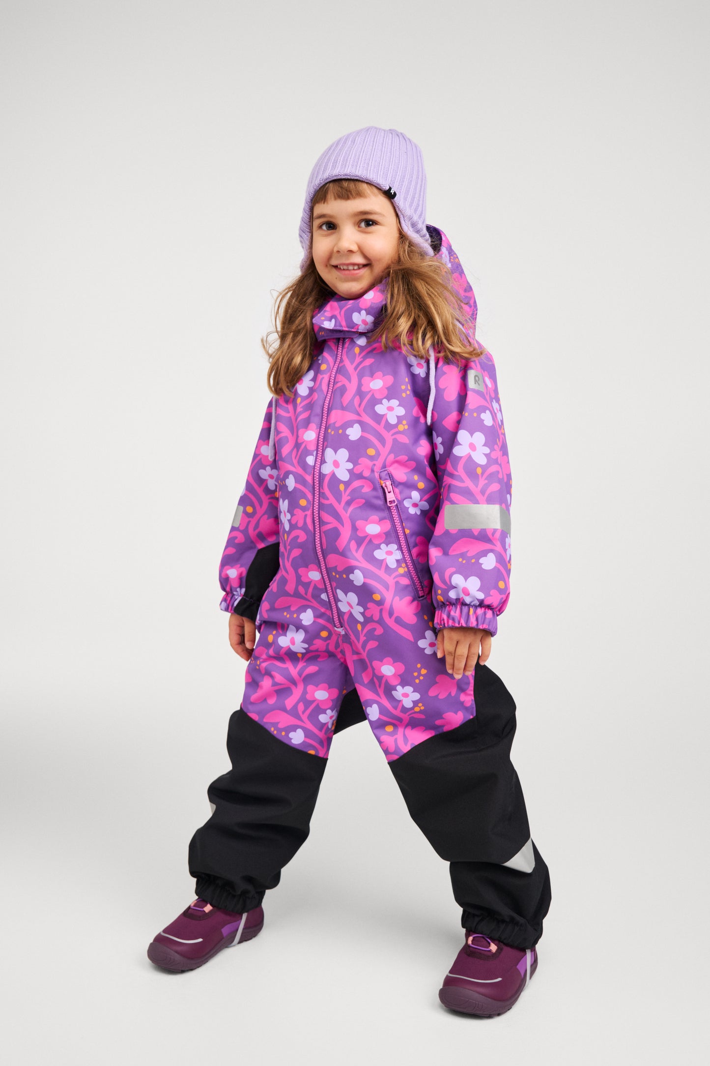 Full-body shot of a young girl standing confidently in the Reima Kurikka waterproof winter snowsuit in plum purple floral, wearing a purple beanie and winter boots, fully outfitted for snow.