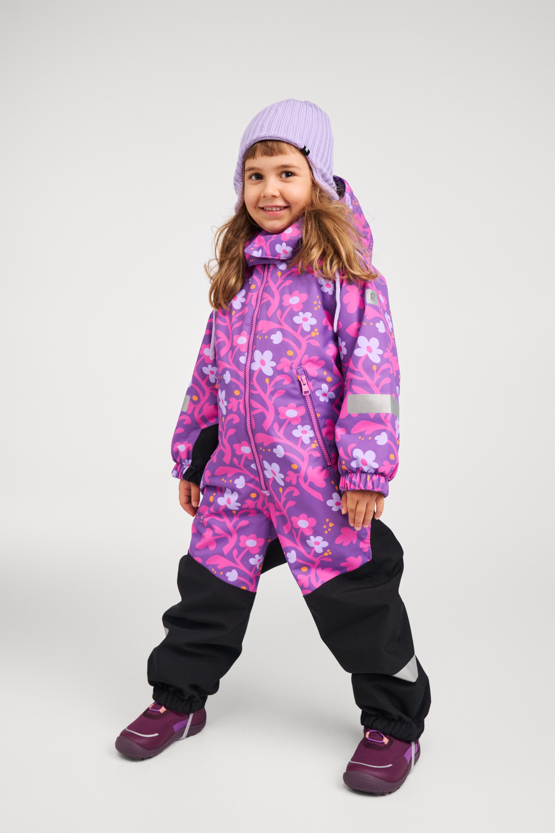 Full-body shot of a young girl standing confidently in the Reima Kurikka waterproof winter snowsuit in plum purple floral, wearing a purple beanie and winter boots, fully outfitted for snow.