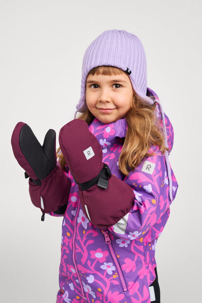 A smiling young girl dressed for winter play wearing the Reima Kurikka waterproof snowsuit in plum purple floral, paired with a light purple ribbed knit beanie and thick maroon Reima winter mittens.
