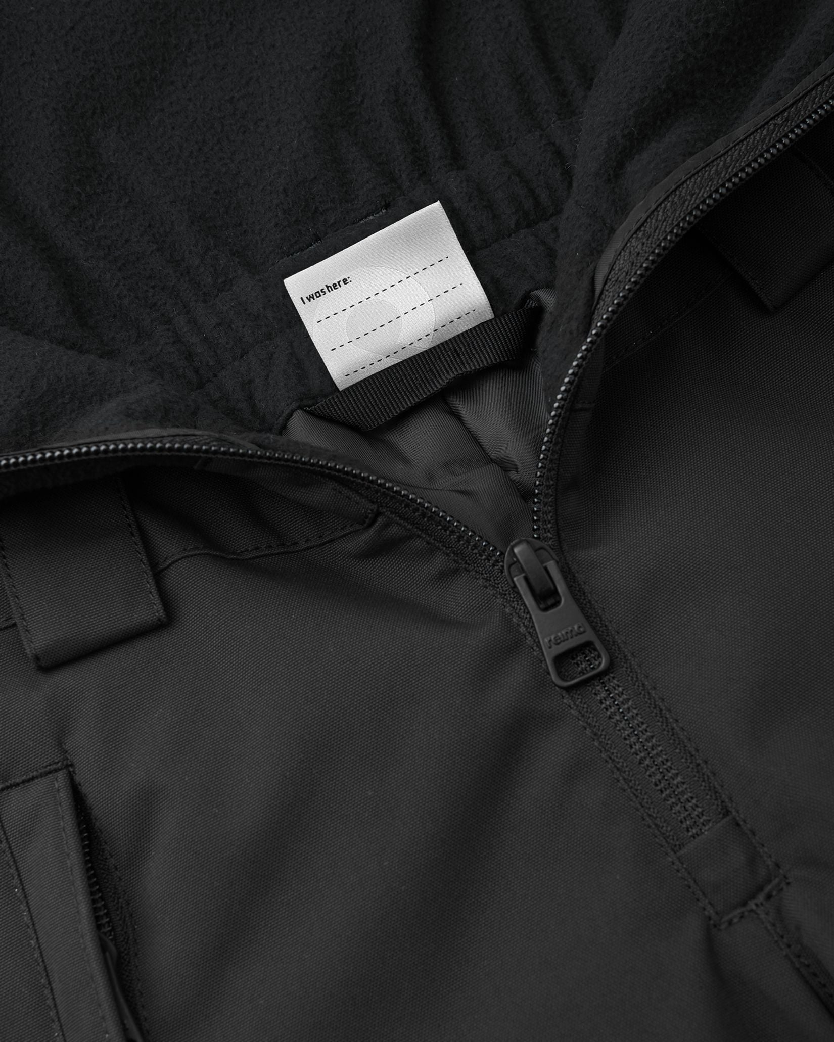 Interior detail view of the black Reima Alppi kids' snow bibs, featuring a customizable blank name tag label inside the soft fleece upper vest for easy identification at ski school or winter camp.