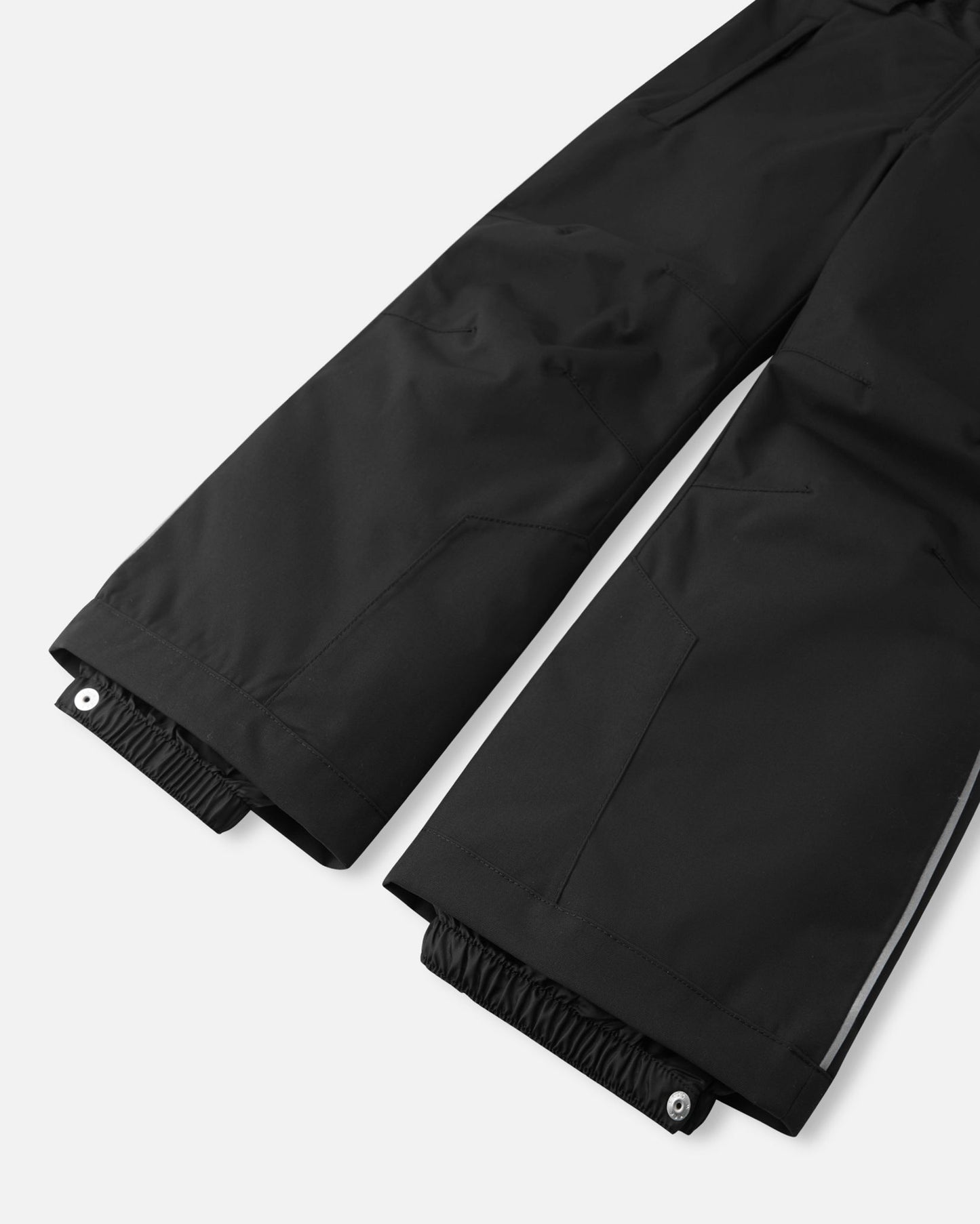Detail view of the lower legs on the black Reima Alppi kids' snow bib pants, emphasizing the elastic inner snow locks with snap closures to keep powder out of boots, reinforced hems for durability, and a vertical safety reflective strip for high visibility on the slopes.