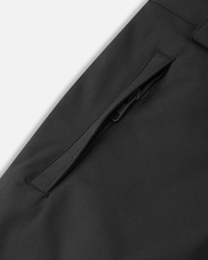 Close-up detail of the leg on the black Reima Alppi kids' snow bibs, highlighting a secure, low-profile zippered pocket designed to safely store small items or a ski pass on the mountain.