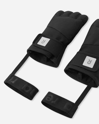 Angled flat lay view of the black Reima Hanskas children's ski gloves, emphasizing the open cuffs for easy layering, adjustable hook-and-loop wrist straps, and the attached elastic safety wrist leashes designed to prevent dropped gloves on the chairlift.