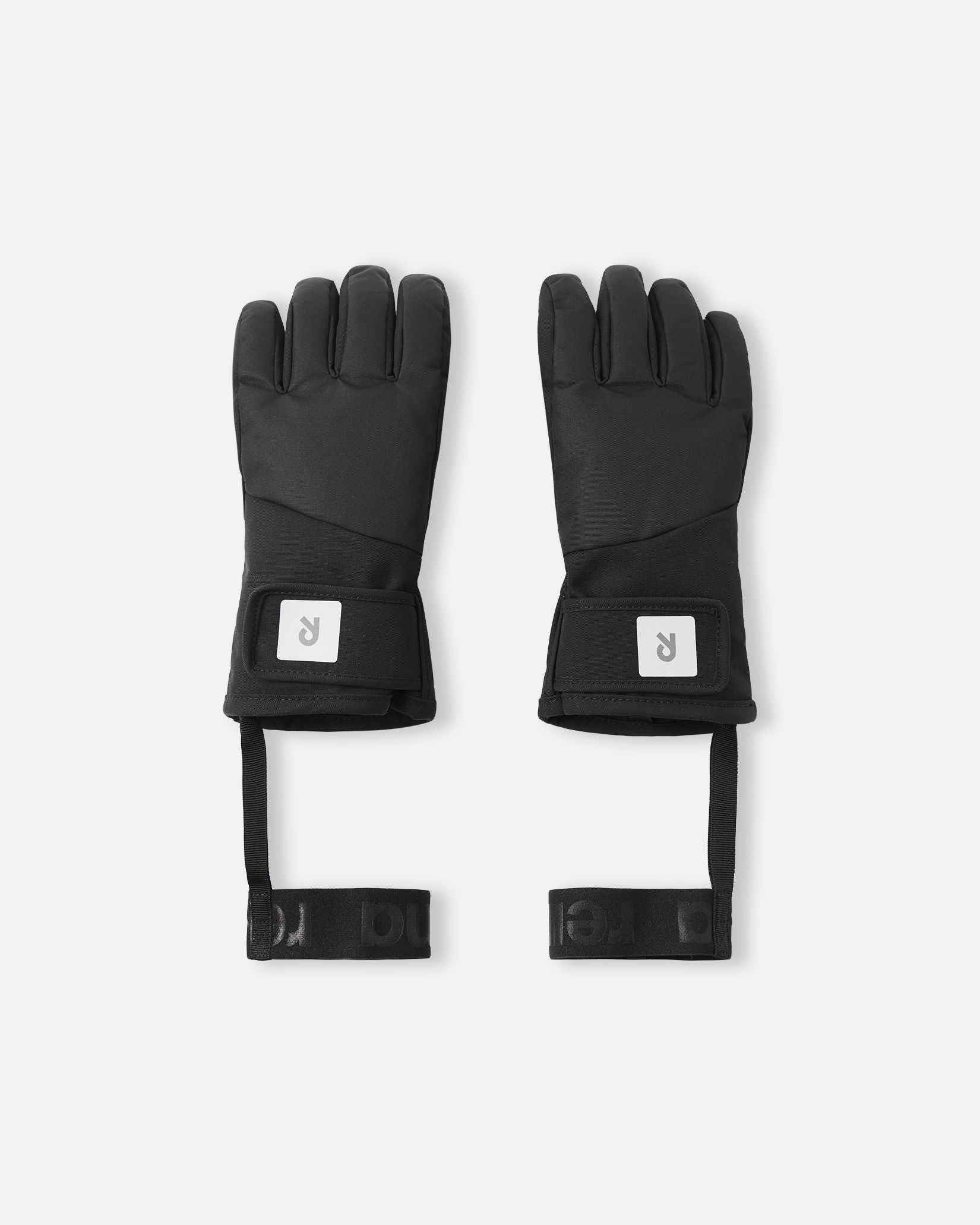 Flat lay view of the backs of the classic black Reima Hanskas kids' ski gloves. The image highlights the waterproof technical fabric, an easy-to-use hook-and-loop adjustable wrist strap for a secure fit, and attached elastic safety wrist leashes.