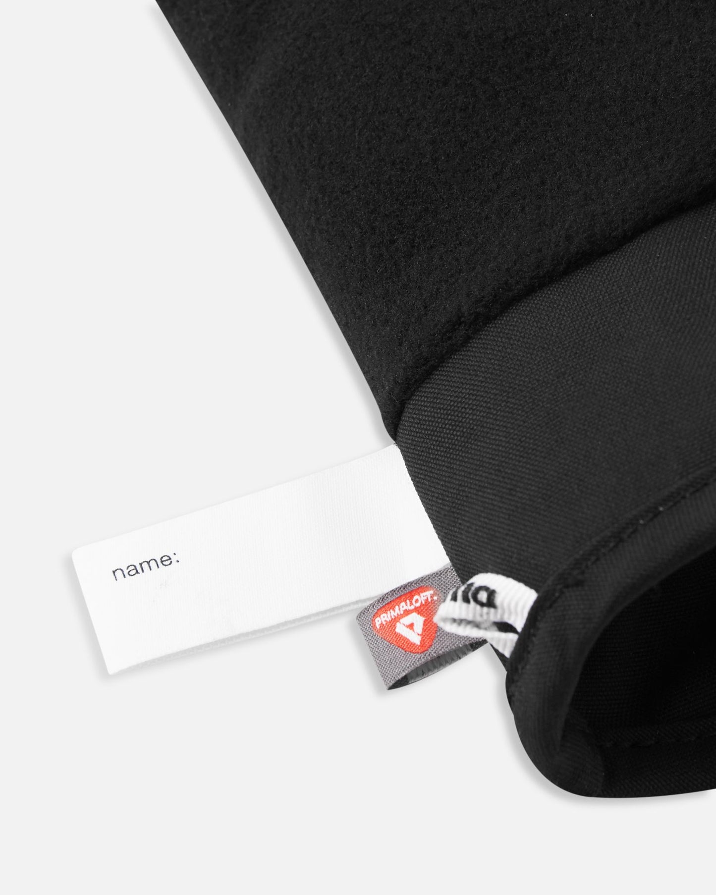 Close-up alternate detail of the interior of the black Reima Hanskas kids' ski gloves, clearly displaying a blank white customizable name tag and the recycled PrimaLoft insulation label attached to the cozy fleece lining.