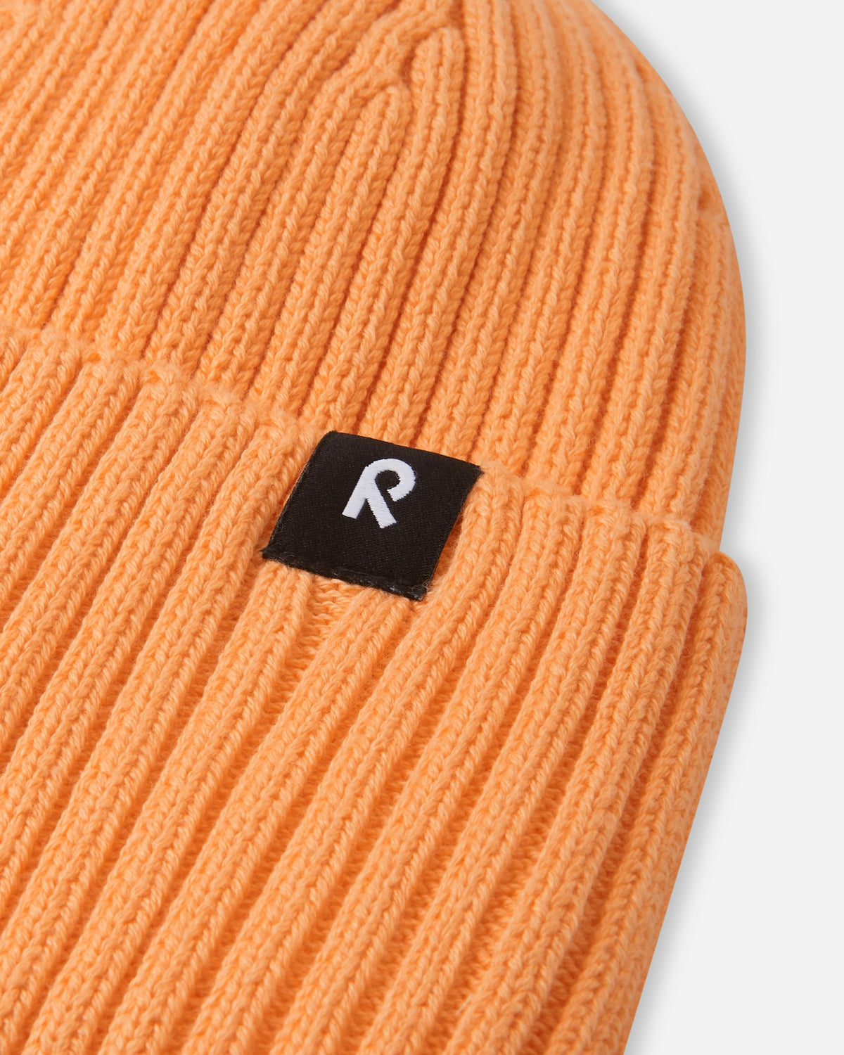 Close-up texture detail of the soft organic cotton ribbed knit on the apricot Reima Hattara kids' beanie, showing the contrast black-and-white logo tag.