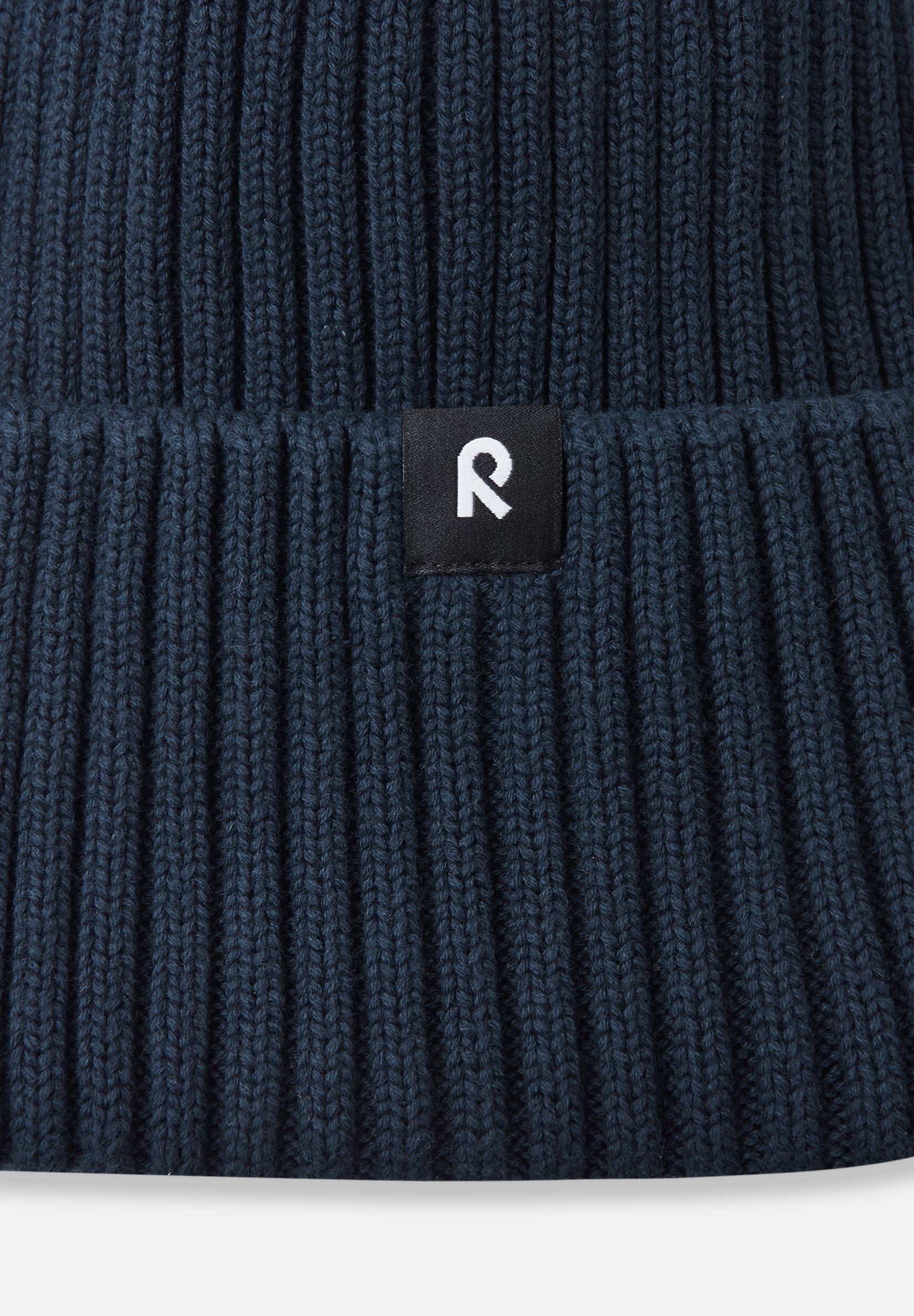 Macro close-up of the soft, breathable organic cotton ribbed knit fabric used on the navy Reima Hattara kids' beanie, featuring the branded logo patch.