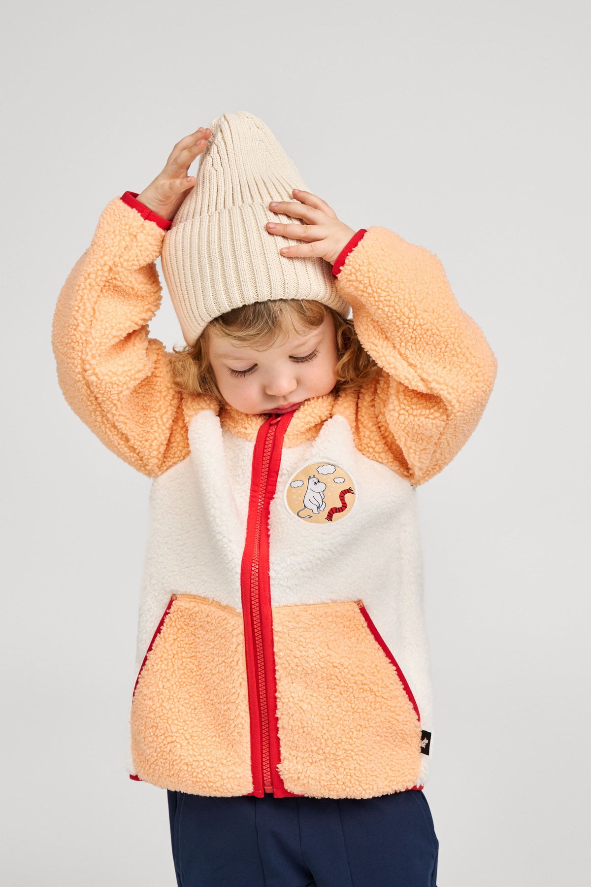 Lifestyle shot of a young child wearing the birch beige Reima Hattara kids' organic cotton beanie paired with a cozy sherpa fleece jacket, demonstrating its comfortable everyday fall wear fit.