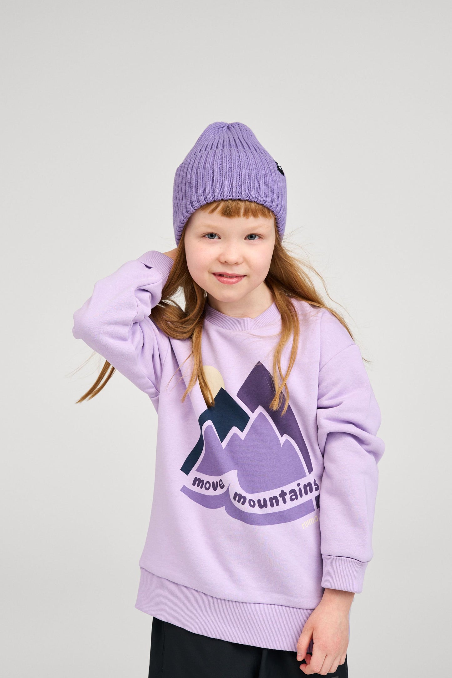 Lifestyle shot of a young child wearing the misty violet Reima Hattara kids' organic cotton beanie paired with a matching purple graphic sweatshirt, demonstrating its everyday wear fit.