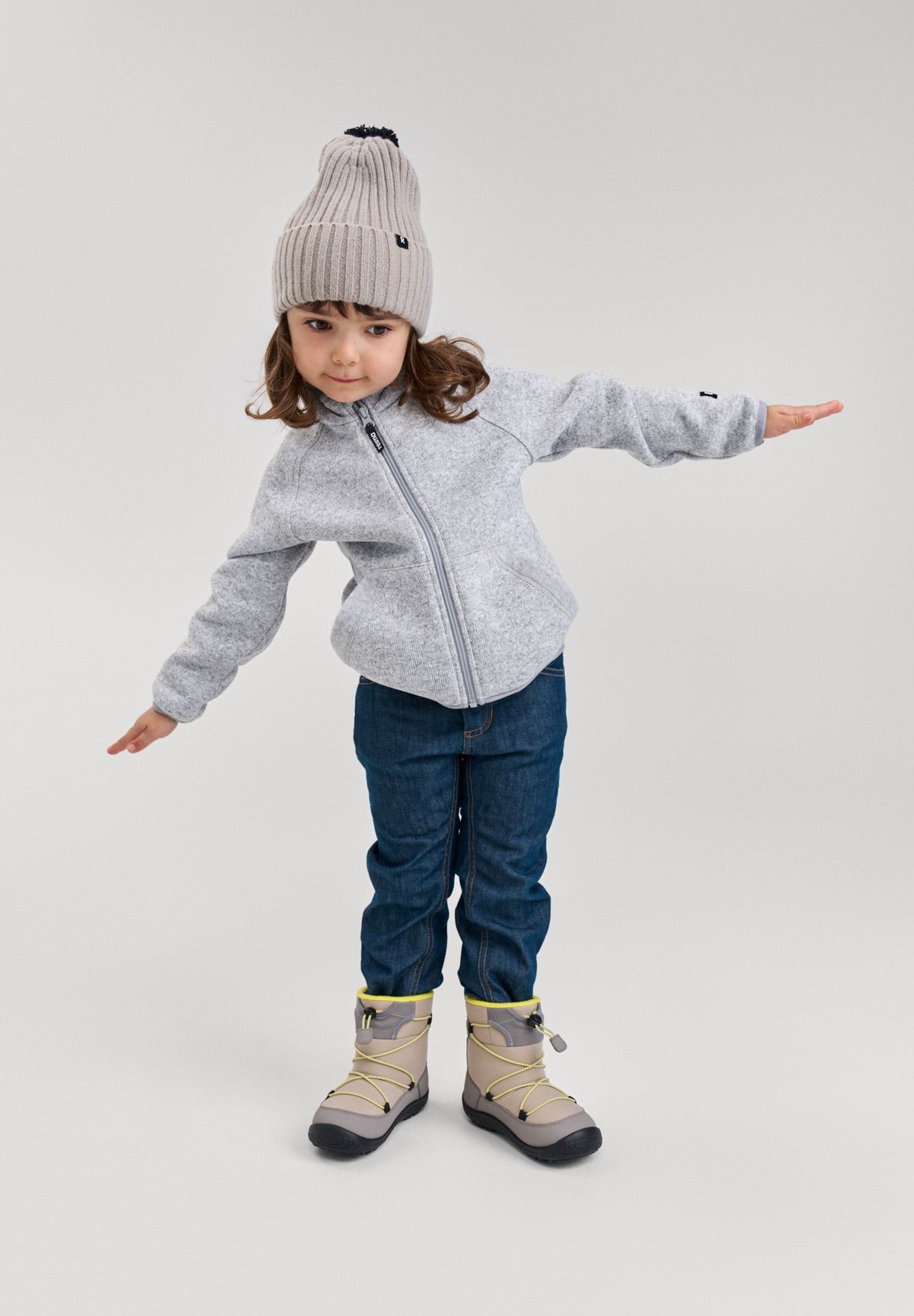 Lifestyle photo of a child modeling the melange grey Reima Hopper kids' knit fleece jacket with arms outstretched, styled with a winter beanie, jeans, and boots to showcase active everyday wear and fit.