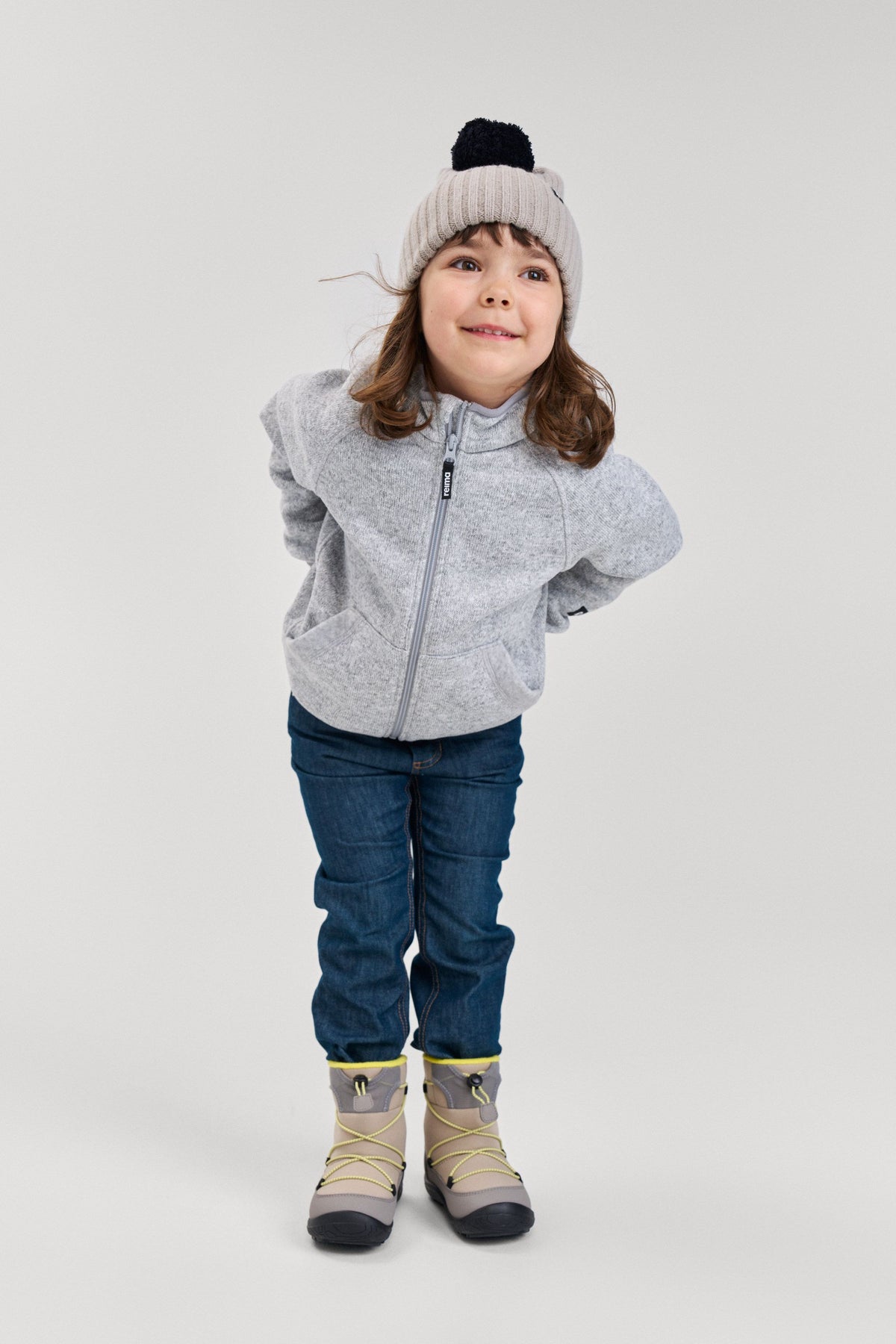 Lifestyle shot of a young child wearing the melange grey Reima Hopper kids' knit fleece jacket paired with jeans and winter boots, demonstrating its versatile and comfortable everyday outdoor fit.
