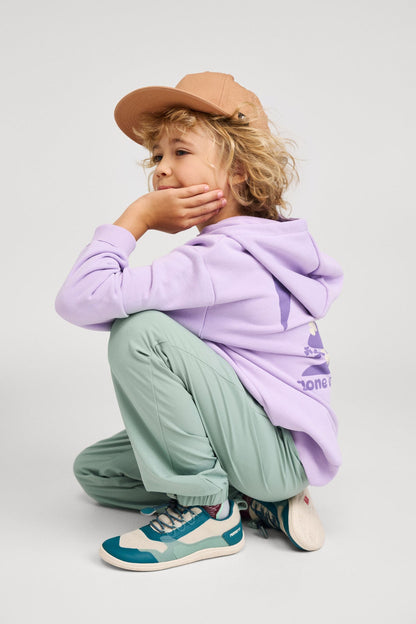 Lifestyle shot of a child wearing the blooming lilac Reima Huppis organic cotton hoodie in a crouching pose, paired with green outdoor pants, a tan cap, and sneakers, demonstrating its flexible, active fit for everyday play.