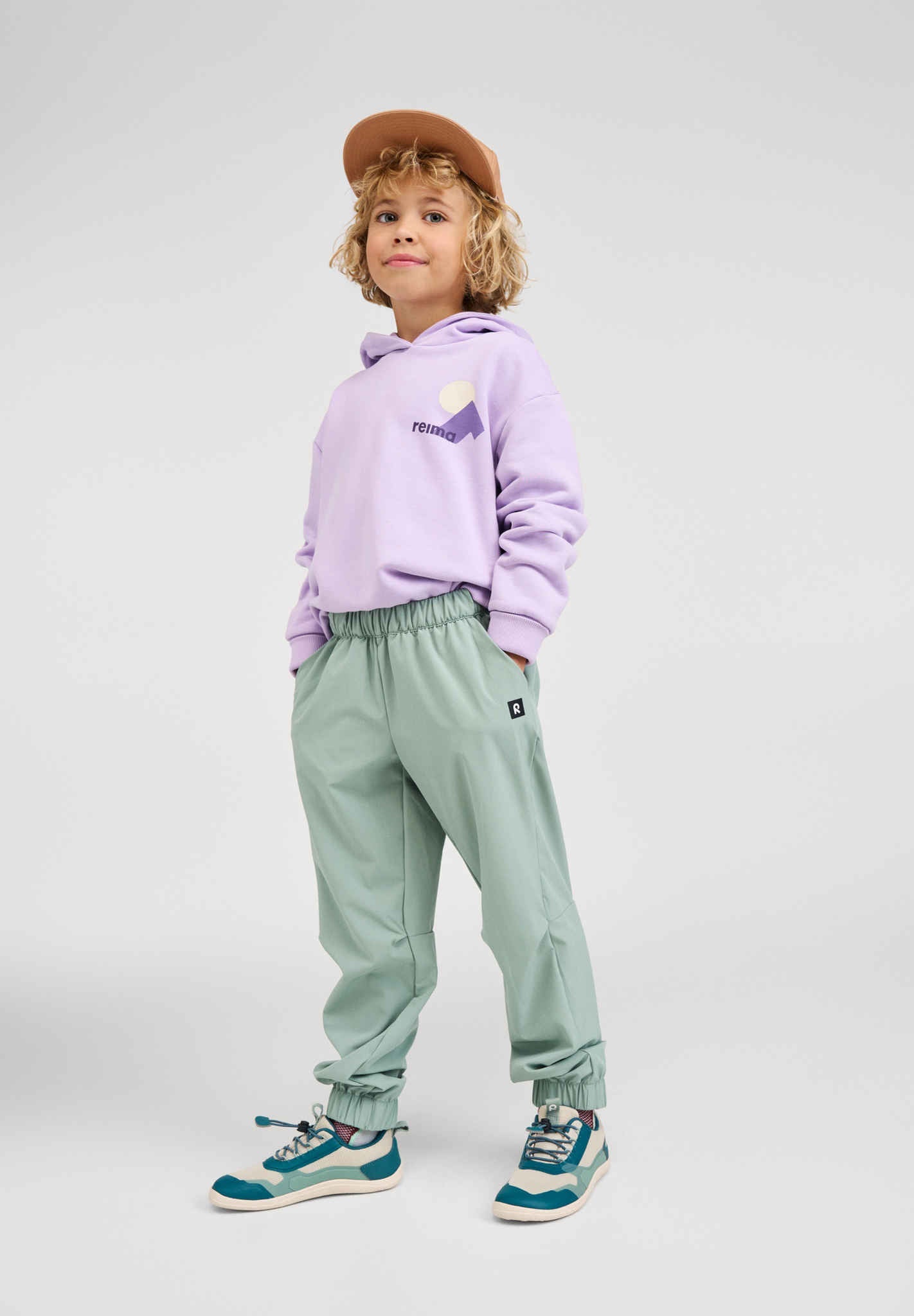 Full-length lifestyle shot of a child modeling the blooming lilac Reima Huppis organic cotton hoodie paired with green outdoor pants, a cap, and sneakers, highlighting versatile kids' apparel styling.