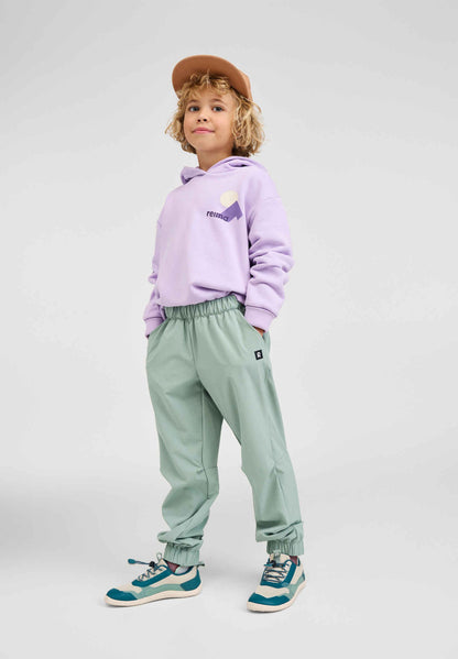 Full-length lifestyle shot of a child modeling the blooming lilac Reima Huppis organic cotton hoodie paired with green outdoor pants, a cap, and sneakers, highlighting versatile kids' apparel styling.