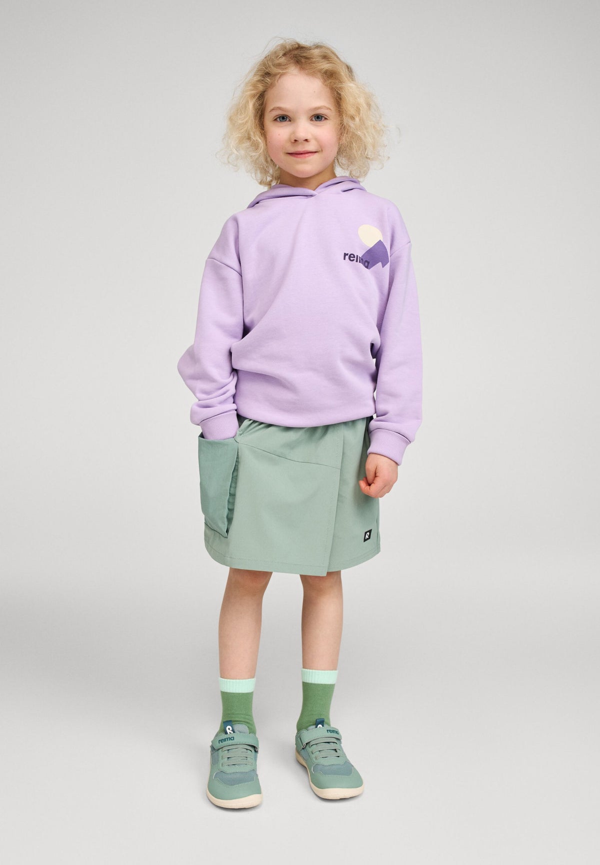Lifestyle photo of a young child wearing the Reima Huppis organic cotton hoodie in blooming lilac featuring a chest graphic logo, styled with a green skirt and sneakers for a casual everyday look.