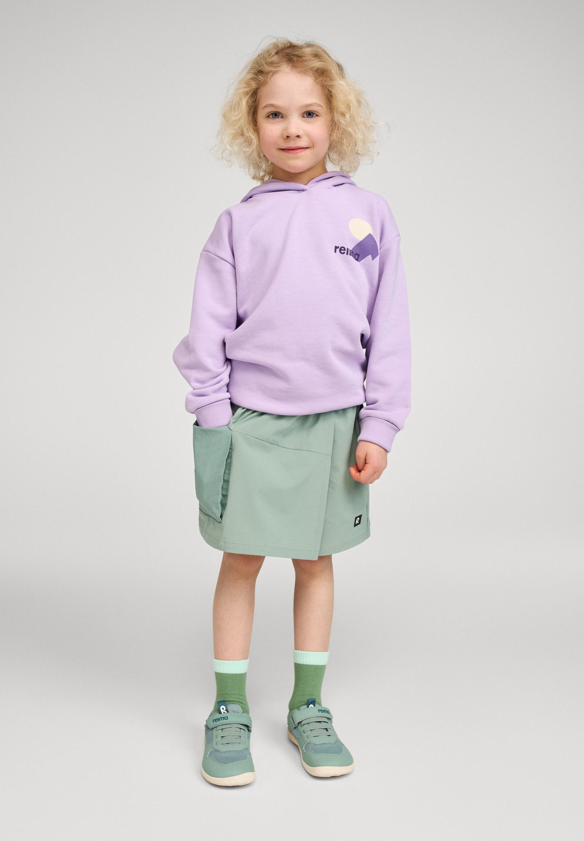 Lifestyle photo of a young child wearing the Reima Huppis organic cotton hoodie in blooming lilac featuring a chest graphic logo, styled with a green skirt and sneakers for a casual everyday look.
