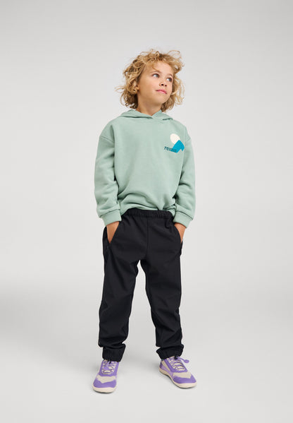 Lifestyle shot of a child wearing the Reima Huppis kids' stone green organic cotton hoodie paired with dark outdoor pants, demonstrating the relaxed, stylish fit ideal for school or trail.