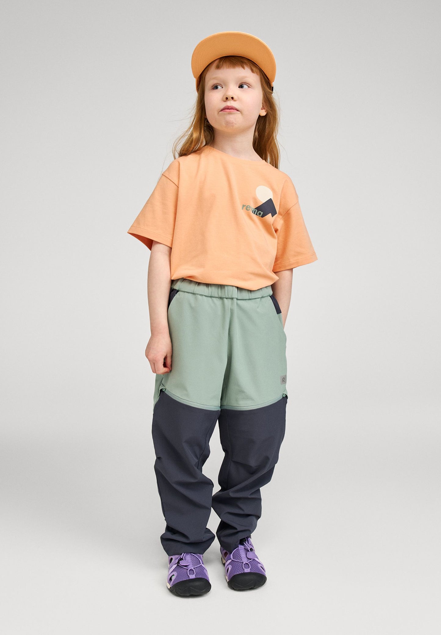 Lifestyle shot of a young girl wearing the apricot Reima Ikioma kids' UPF 50+ sunproof t-shirt paired with green and navy outdoor pants, demonstrating its comfortable, active fit for summer adventures.