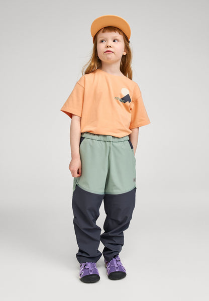 Lifestyle shot of a young girl wearing the apricot Reima Ikioma kids' UPF 50+ sunproof t-shirt paired with green and navy outdoor pants, demonstrating its comfortable, active fit for summer adventures.