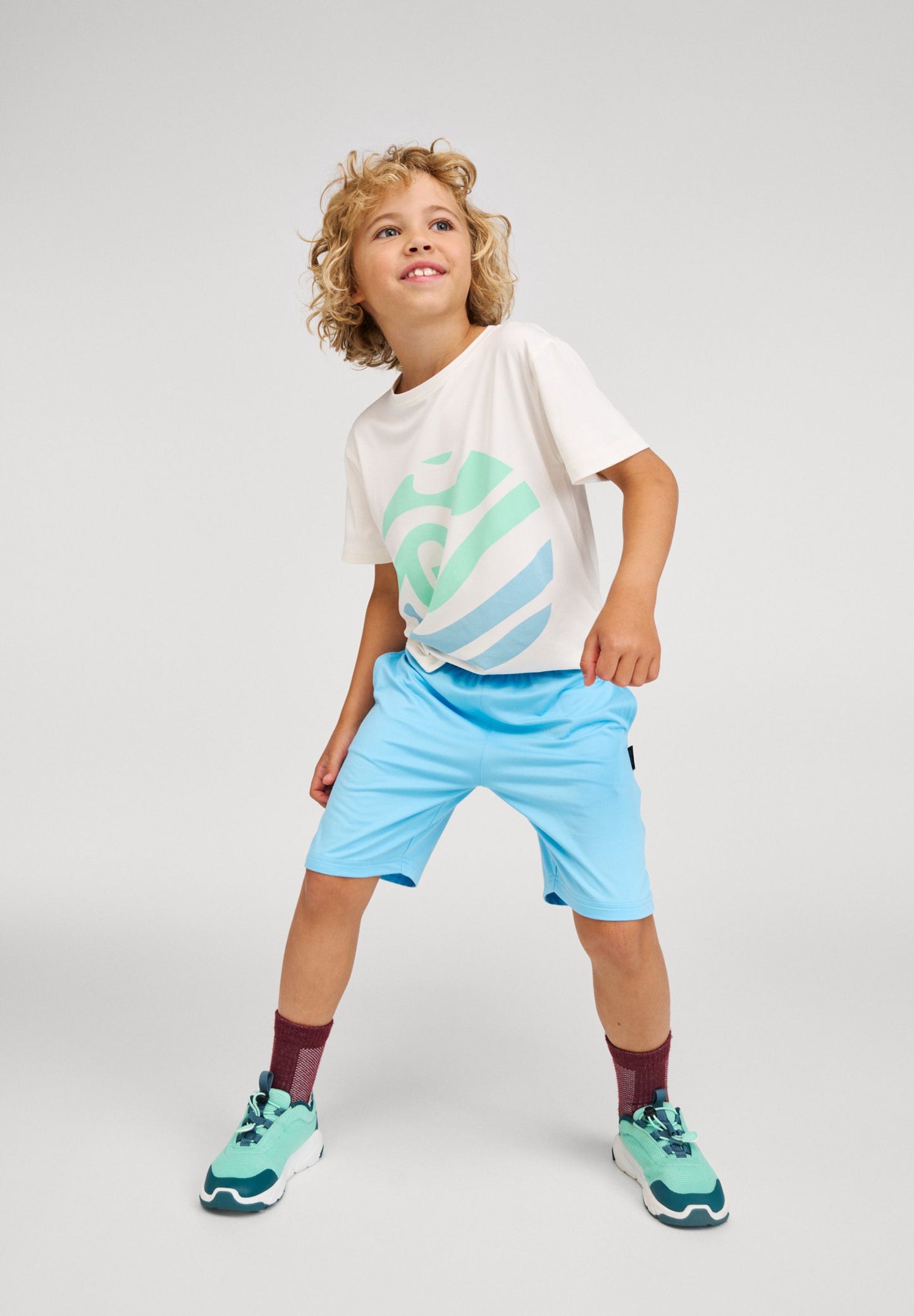 A detailed lifestyle photo of a smiling young boy with curly blonde hair wearing light blue (Fresh Blue) Reima Ilmassa shorts and a white graphic Vauhdikas t-shirt. The child is in an active standing pose against a grey studio background, looking up and to the right, and is also wearing maroon socks and teal-and-white sneakers.