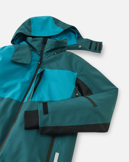 Angled close-up of the dark teal Reima Jatuni kids' ski jacket, demonstrating the safe detachable hood with snap buttons, the robust main front zipper, and a water-repellent zippered chest pocket.