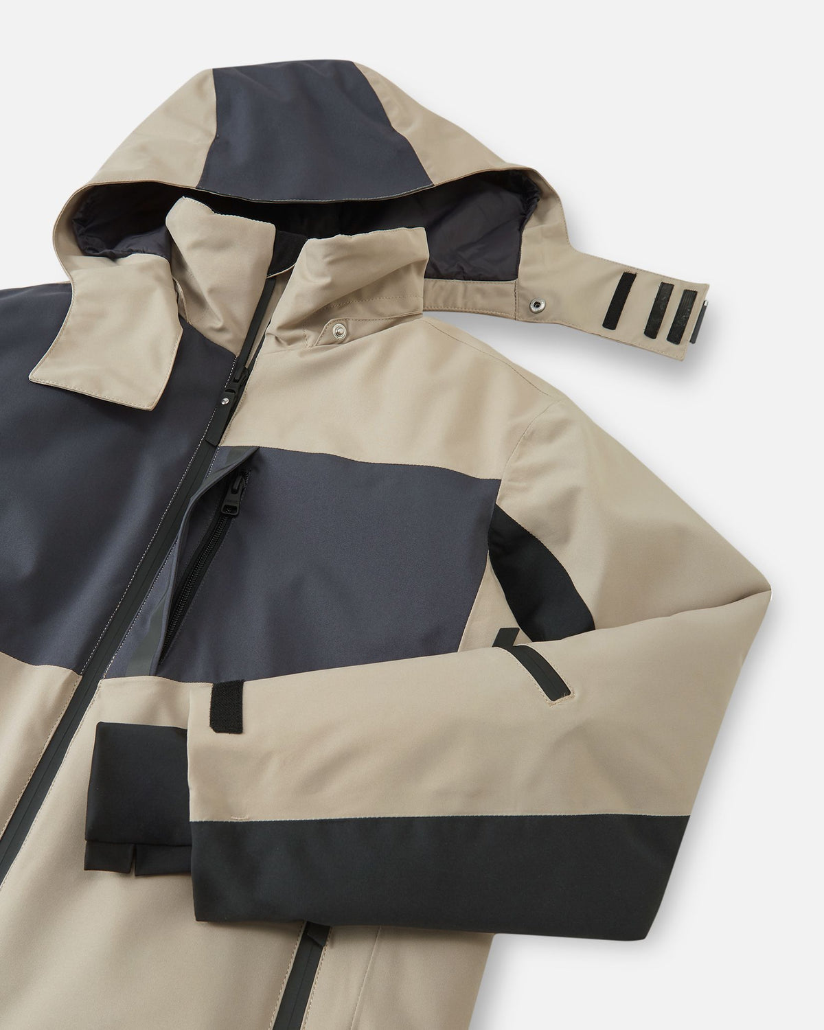 Angled close-up of the earthy beige Reima Jatuni kids' ski jacket, demonstrating the safe detachable hood with snap buttons, the main front zipper, and a water-repellent zippered chest pocket.