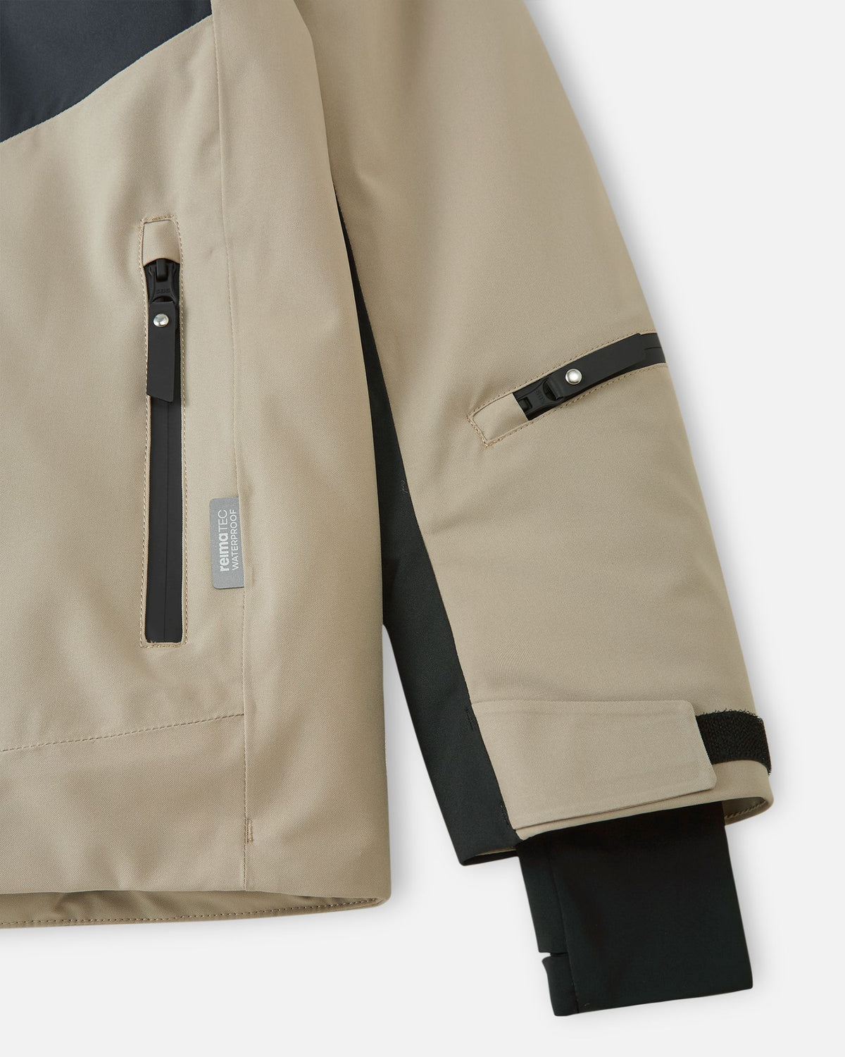 Detail of the sleeve on the earthy beige Reima Jatuni kids' ski jacket, highlighting the zippered ski pass pocket on the forearm, an adjustable hook-and-loop cuff, and a stretchy inner Lycra cuff with a thumbhole.