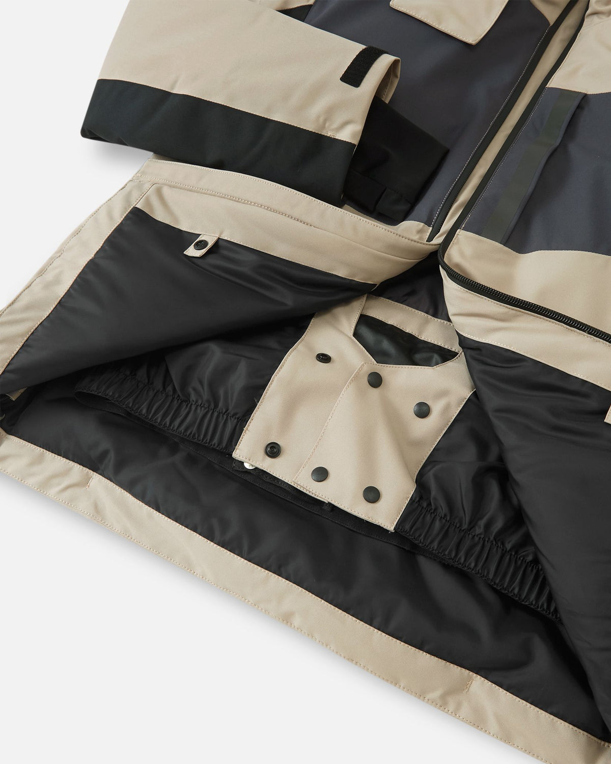 Interior view of the earthy beige Reima Jatuni kids' ski jacket, revealing the built-in adjustable snow skirt with snap buttons designed to keep snow out during alpine skiing and snowboarding.
