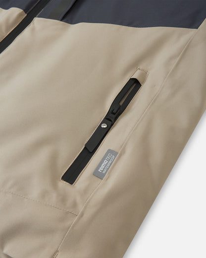 Close-up of a zippered hand pocket on the earthy beige Reima Jatuni kids' ski jacket, featuring a Reimatec waterproof tag indicating premium weather protection and durability.
