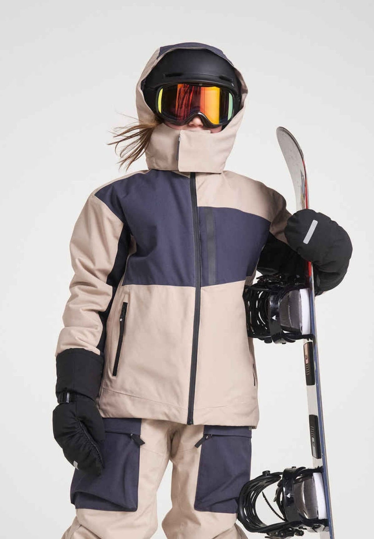 A child holding a snowboard wearing the Reima Jatuni kids' ski jacket in Earthly Beige with a black helmet and goggles.