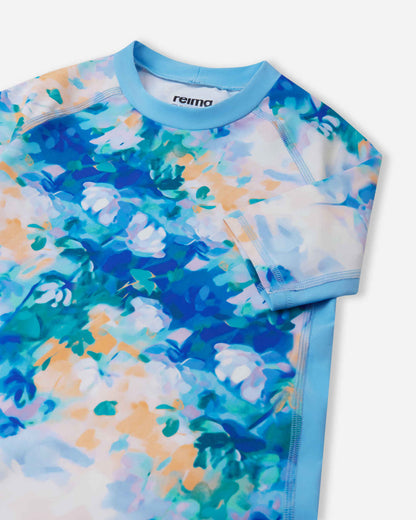 Close-up detail of the Reima Joonia girls' sunproof rash guard in fresh blue, highlighting the protective stretch-knit crew collar, durable flatlock seams, and stylish pastel floral pattern.