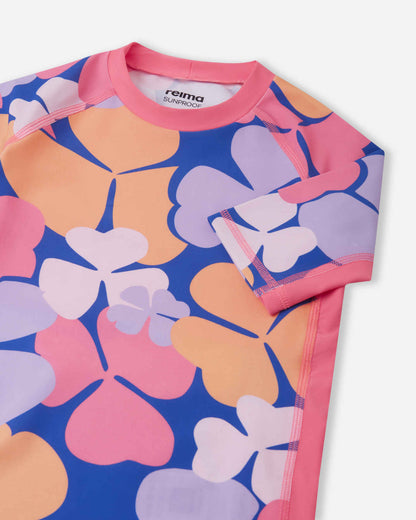 Detailed close-up of the Reima Joonia girls' soft coral sunproof rash guard, focusing on the solid pink collar and short sleeve, smooth flatlock stitching, and the Reima Sunproof label inside the neck.