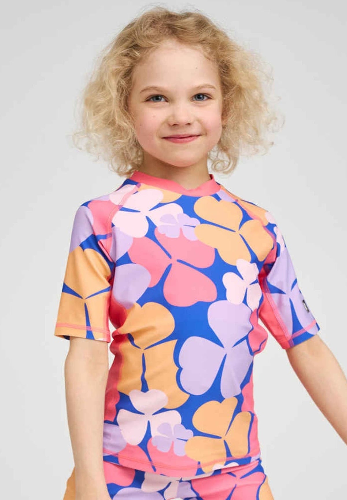 Lifestyle shot of a smiling young girl wearing the soft coral Reima Joonia UPF 50+ sunproof rash guard. The short-sleeve swim shirt features a vibrant oversized floral print and solid pink raglan sleeves, demonstrating its comfortable, stretchy fit designed for active summer swimming and maximum UV protection.