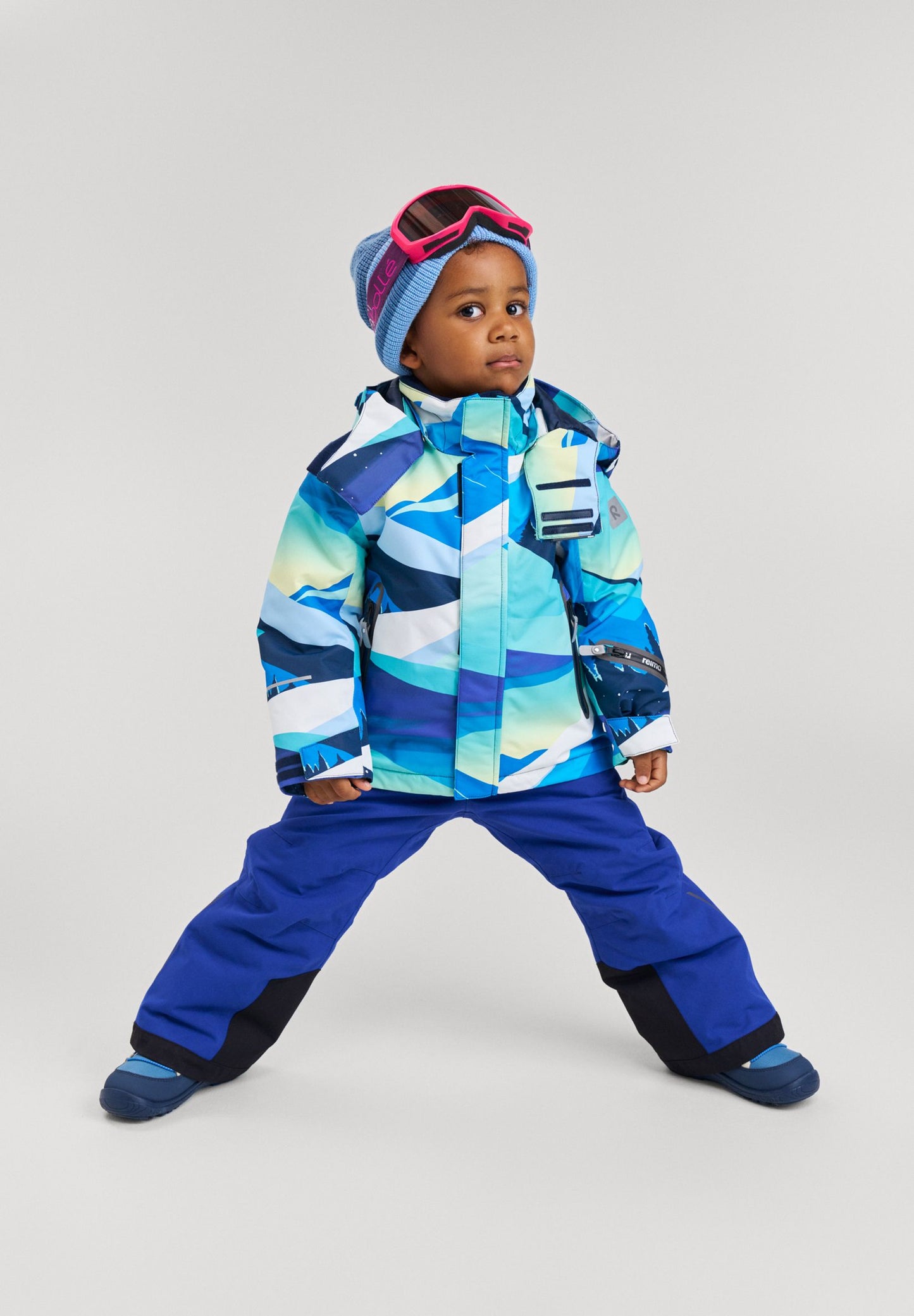 Full-body lifestyle shot of a young child geared up for the mountain in the bright blue Reima Kairala ski jacket, styled with snow pants and ski goggles for ultimate winter protection.