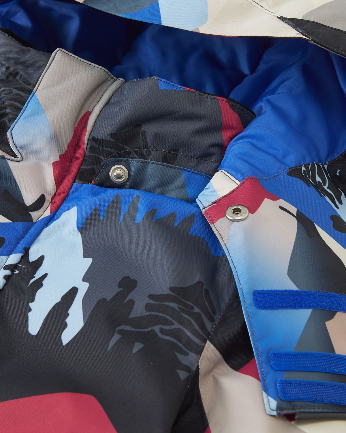Macro detail shot of the collar on the Reima Kairala kids' ski jacket, highlighting the secure metallic snap buttons used to attach the removable safety hood to the soft black mountain patterned fabric.
