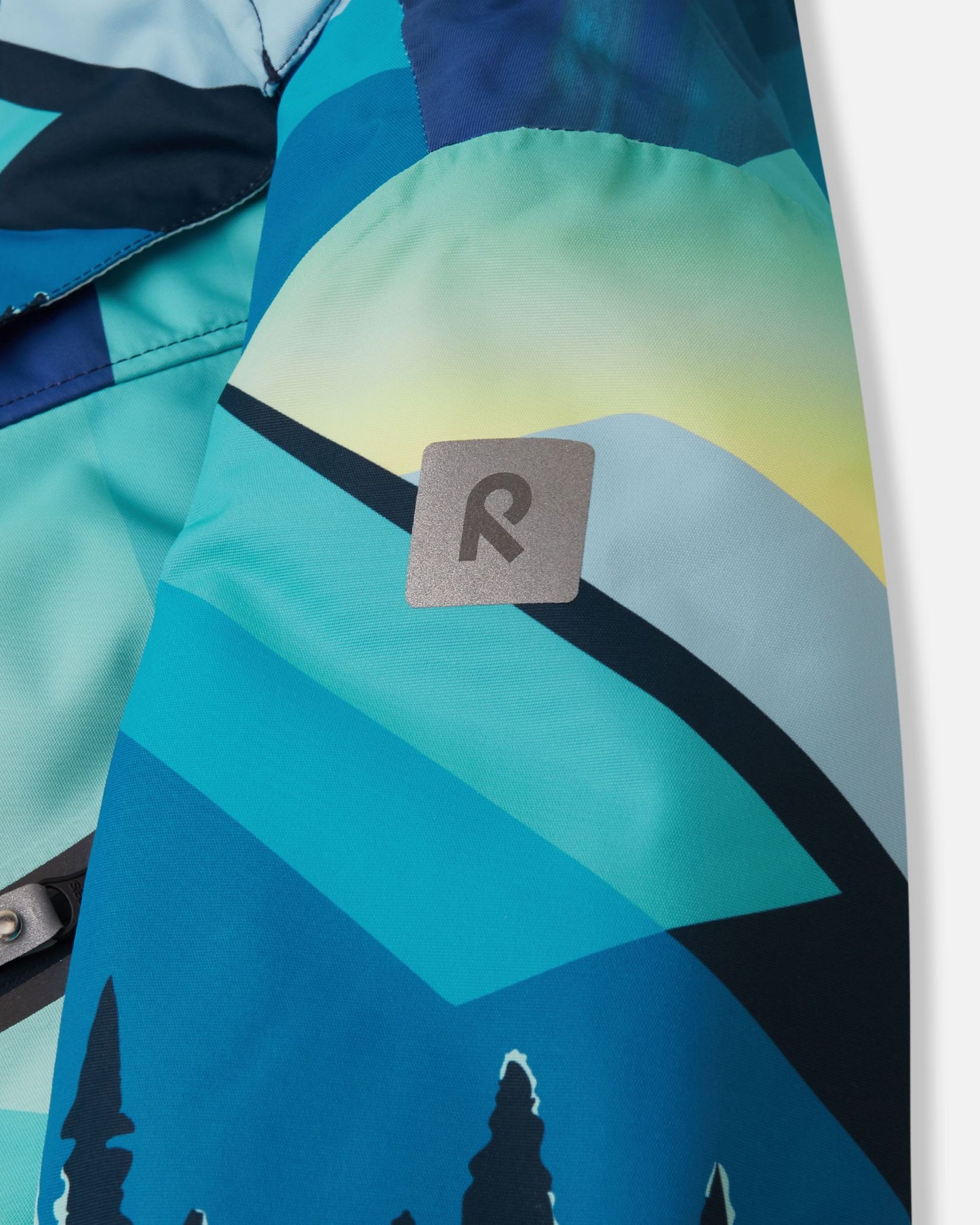 Close-up of the safety-enhancing reflective Reima logo patch on the sleeve of the bright blue Kairala kids' waterproof ski jacket.