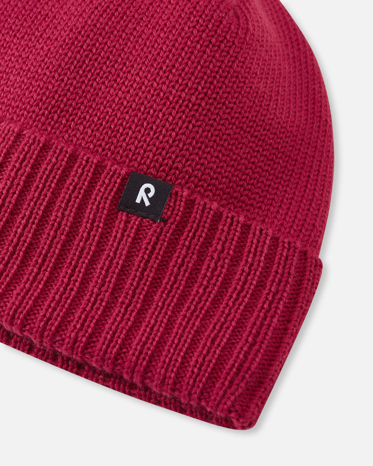 Detailed macro shot of the cranberry red Reima Kalotti kids beanie, emphasizing the high-quality woven merino wool texture, the thick ribbed folding edge for durability, and the small black Reima authenticity logo.