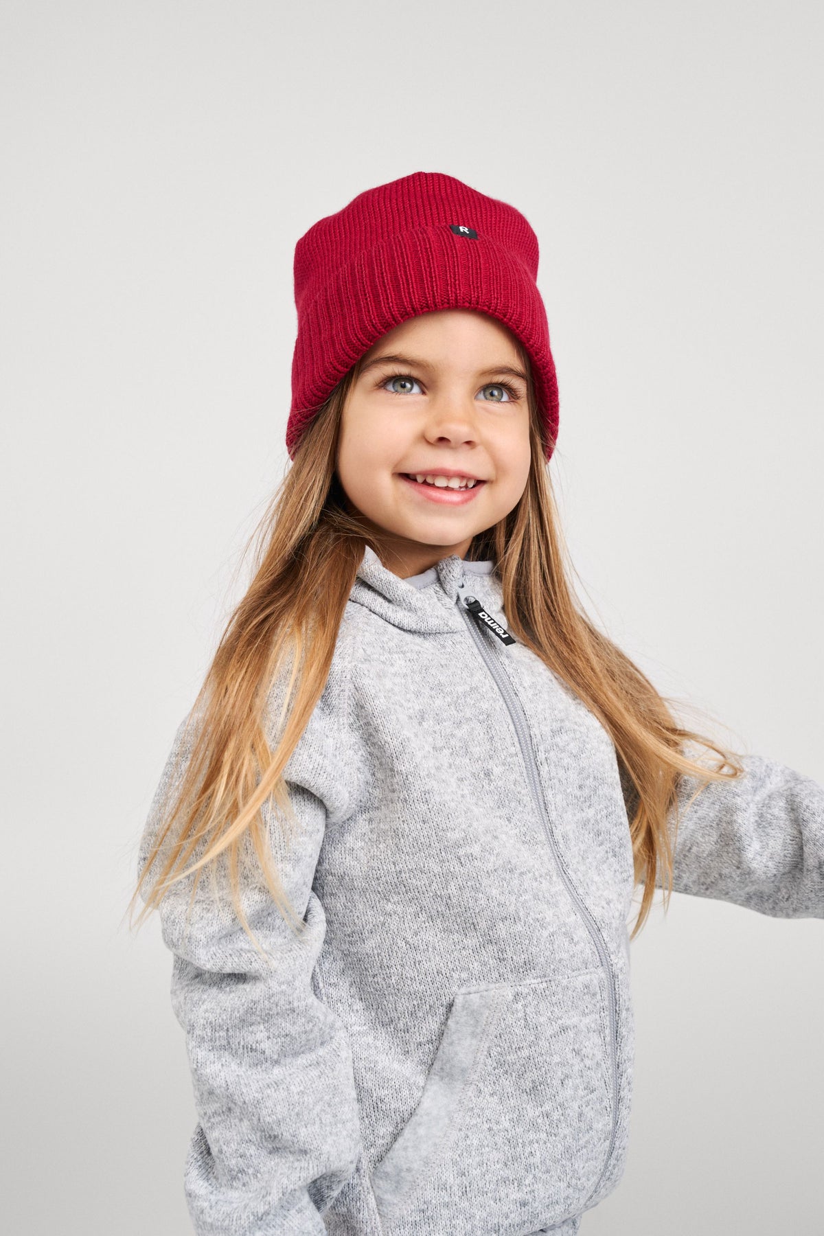 Lifestyle image of a smiling young girl modeling the cranberry red Reima Kalotti kids merino wool beanie, demonstrating how the hat fits snugly over the ears and pairs with a light grey zip-up fleece jacket for fall and winter outdoor wear.