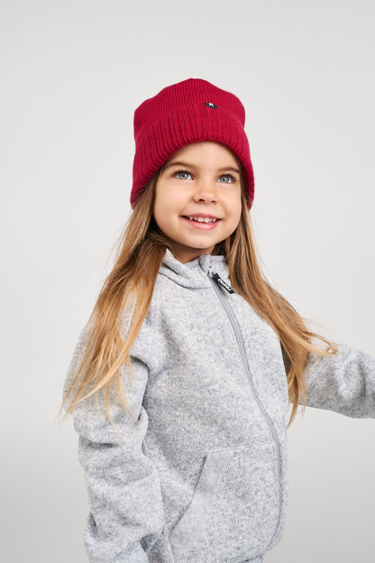 Lifestyle image of a smiling young girl modeling the cranberry red Reima Kalotti kids merino wool beanie, demonstrating how the hat fits snugly over the ears and pairs with a light grey zip-up fleece jacket for fall and winter outdoor wear.