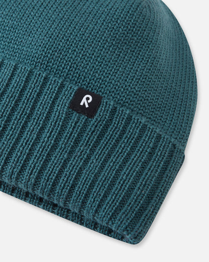 Extreme close-up of the dark teal Reima Kalotti children's winter hat, highlighting the chunky ribbed knit cuff that ensures a snug fit, constructed from breathable merino wool, and finished with the signature black Reima 'R' logo tag.