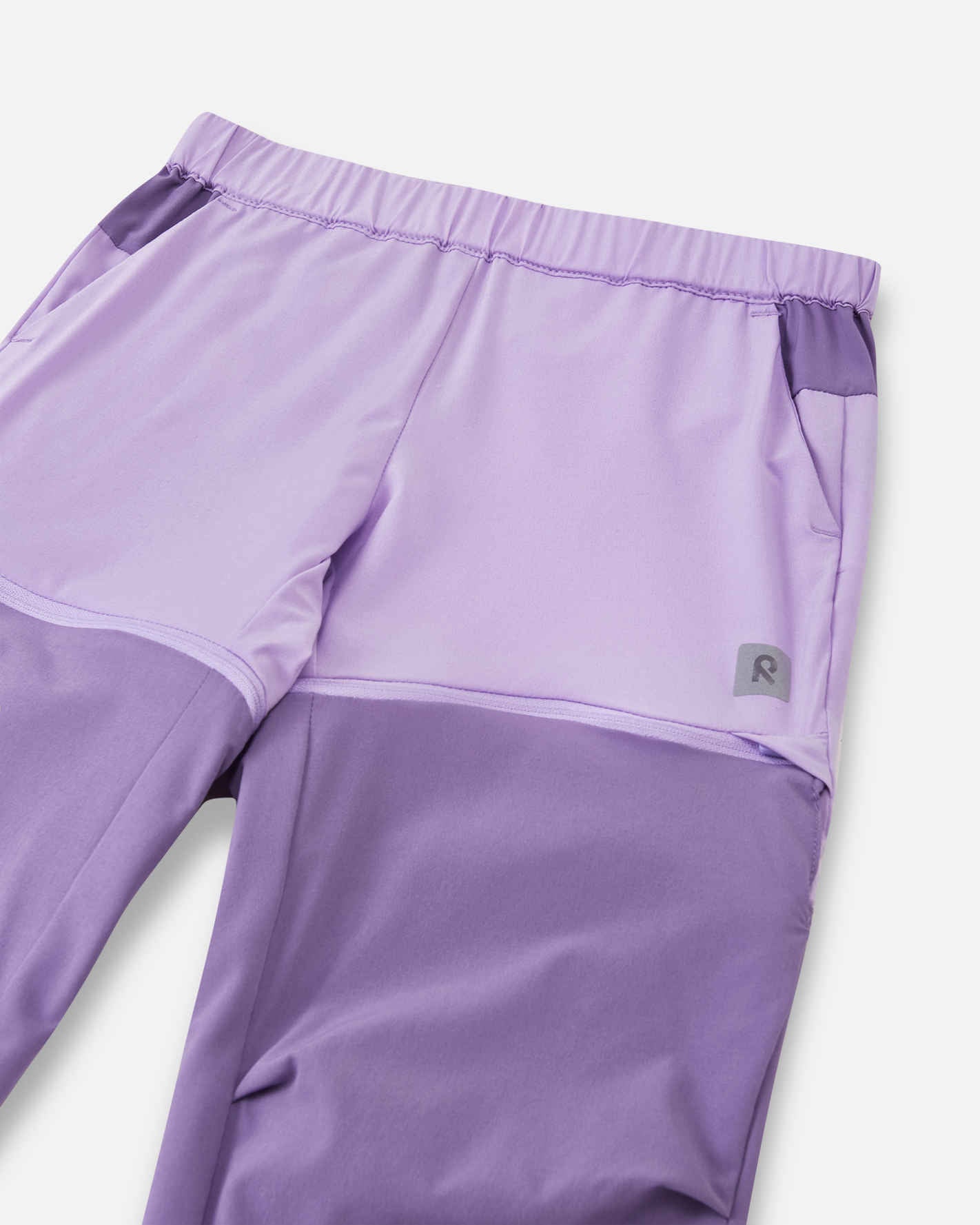 Close-up view of the upper front on the Reima Kaveris kids' blooming lilac hiking pants, highlighting the stretchy elastic waistband, dark purple pocket accents, and a reflective Reima logo.