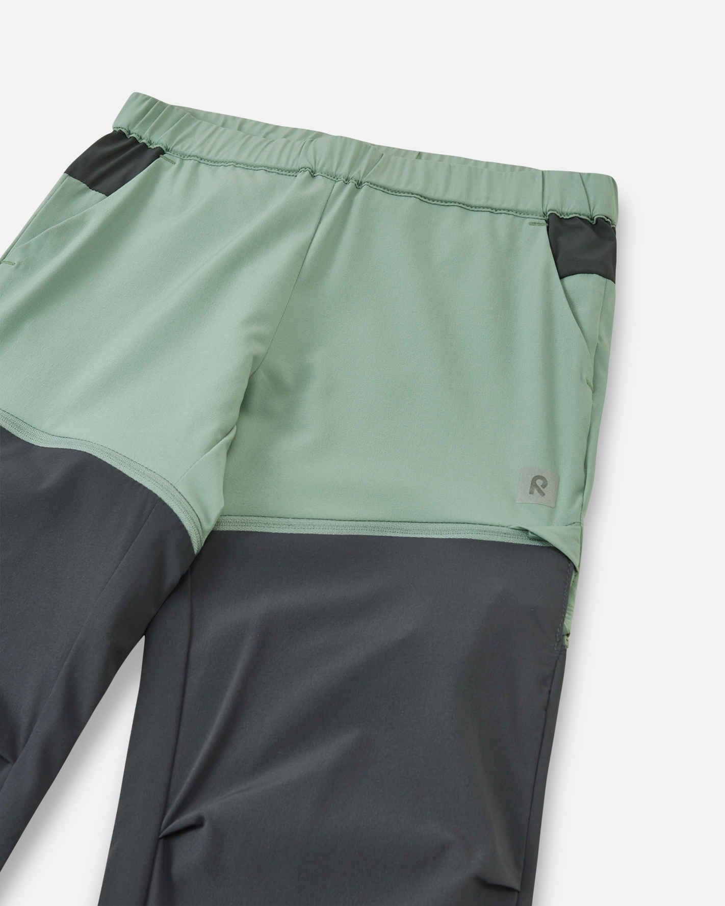 Close-up view of the upper front on the Reima Kaveris kids' stone green hiking pants, highlighting the flexible elastic waistband, dark grey pocket accents, and the small reflective Reima logo.