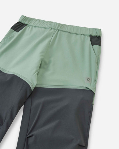 Close-up view of the upper front on the Reima Kaveris kids' stone green hiking pants, highlighting the flexible elastic waistband, dark grey pocket accents, and the small reflective Reima logo.
